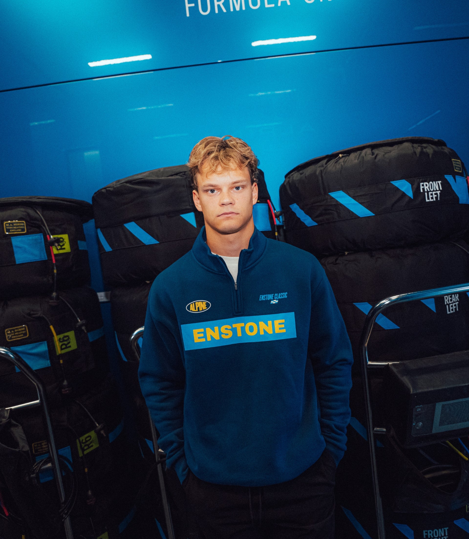 Person wearing a blue Alpine Classic F1 half zip jacket with ENSTONE patch in a gear storage area