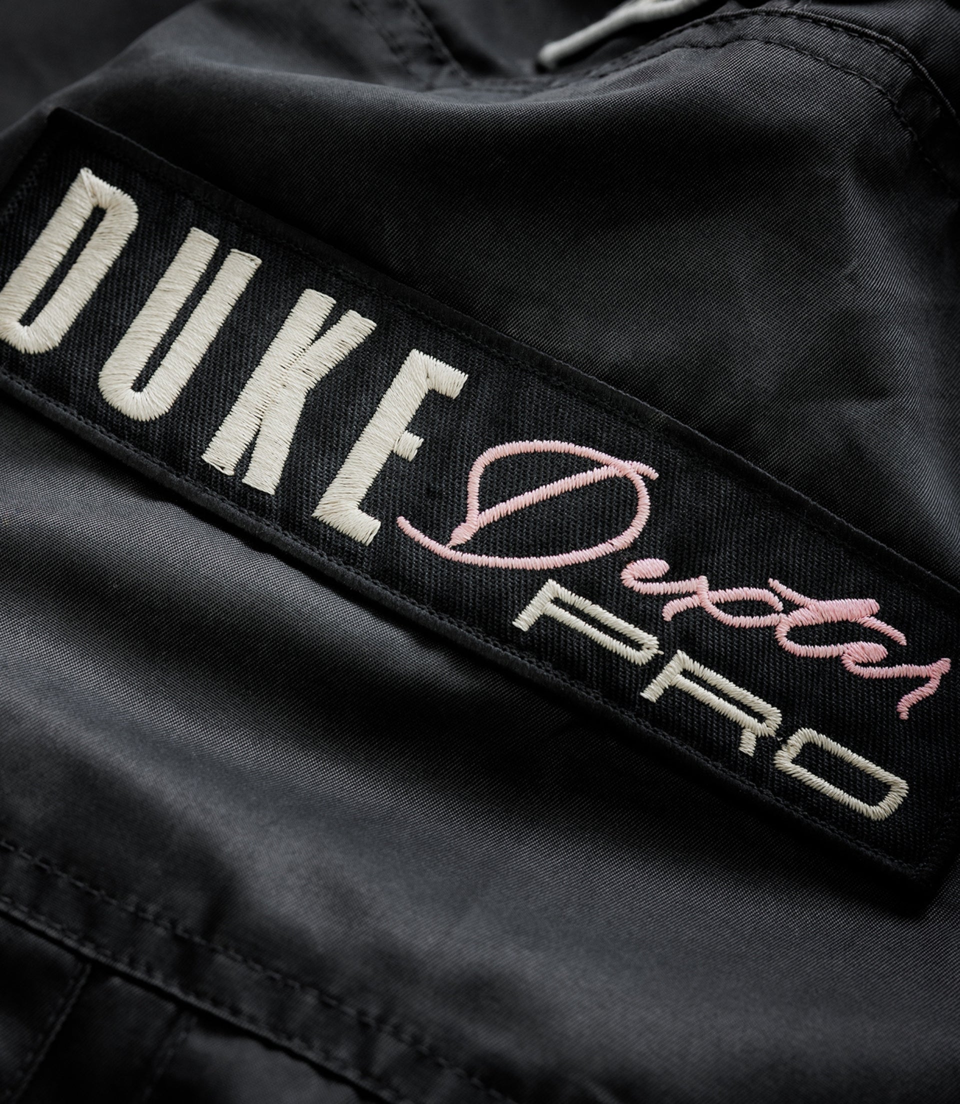 Close-up of black trackside jacket fabric with a white embroidered patch and pink script from the BWT Alpine x Duke & Dexter range