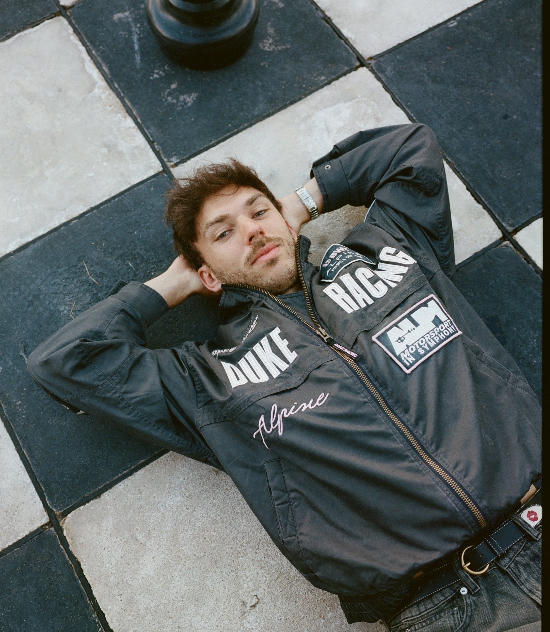 Man lounging on a tiled black and gray pavement wearing a black trackside jacket with large white Duke lettering and racing patches.