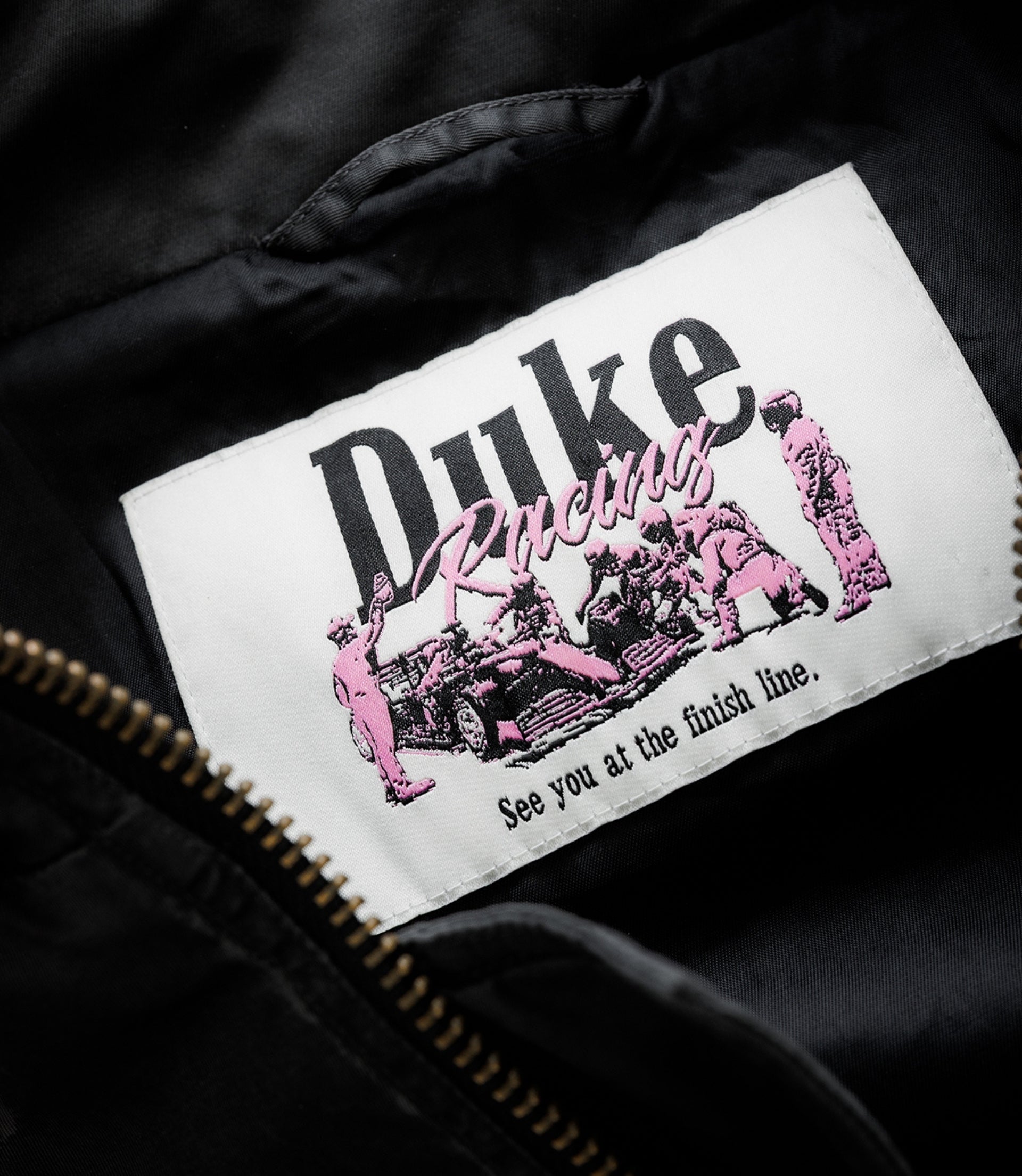White embroidered patch on black fabric showing Duke & Dexter branding with pink racing graphic