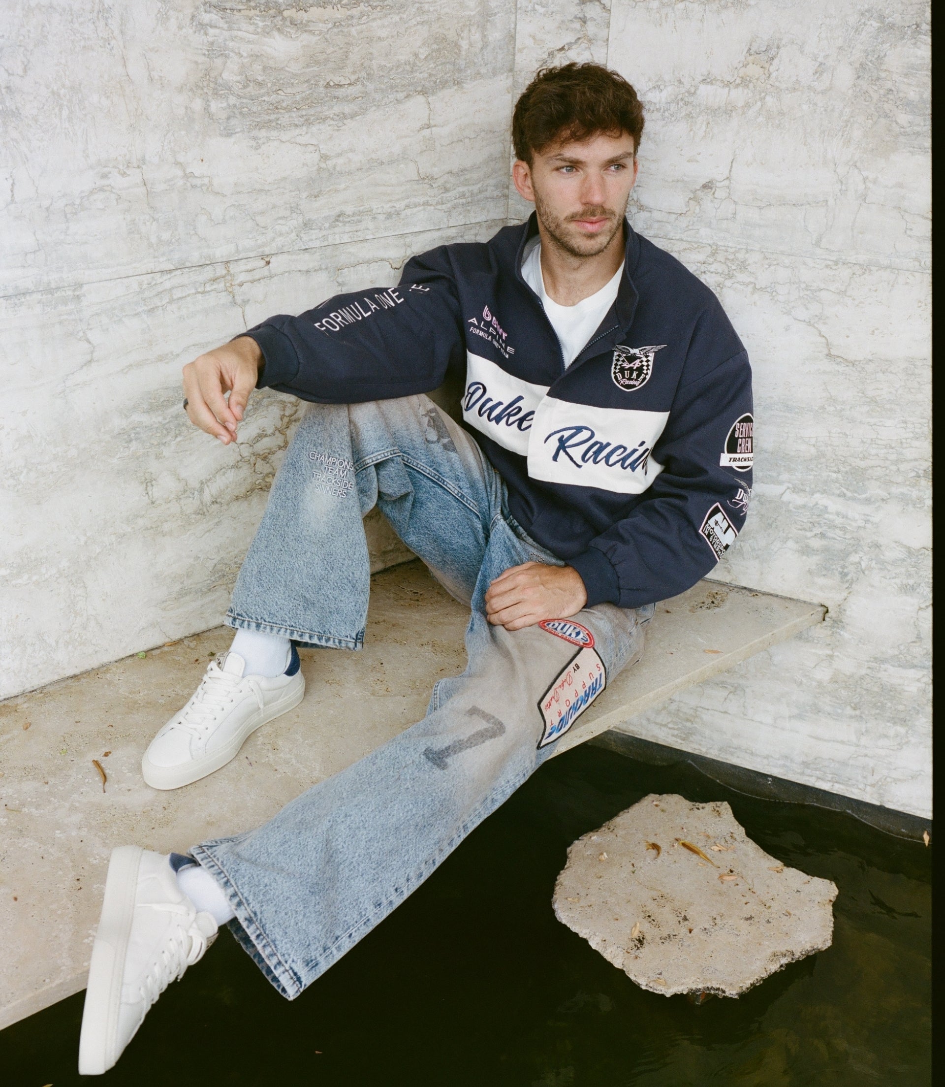 Person wearing navy Duke & Dexter racing jacket with patches sits on stone steps beside a light marble wall