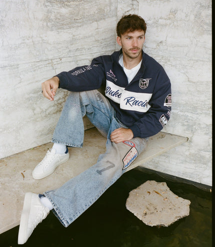 Person wearing navy Duke & Dexter racing jacket with patches sits on stone steps beside a light marble wall