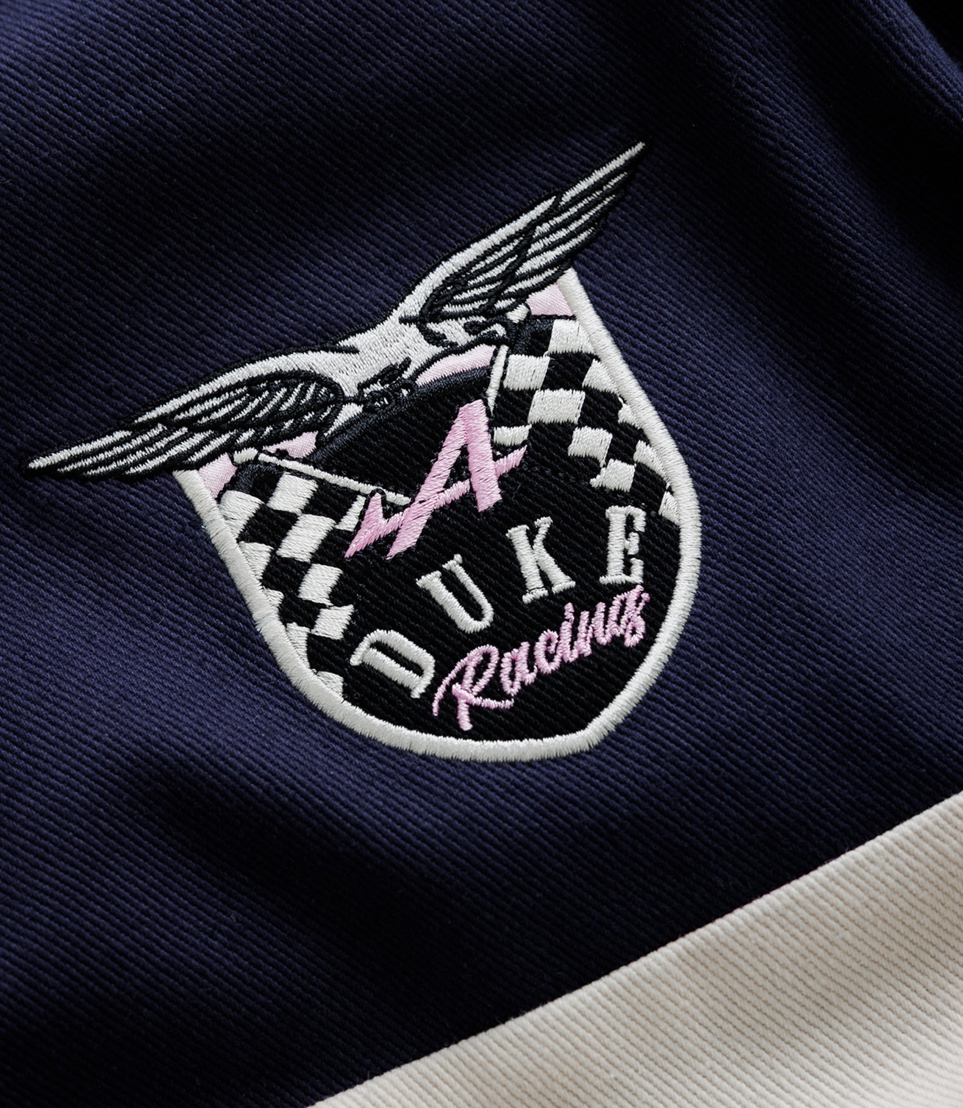 Close-up embroidery of a racing crest on navy fabric showing wings a checkered shield and pink Duke lettering
