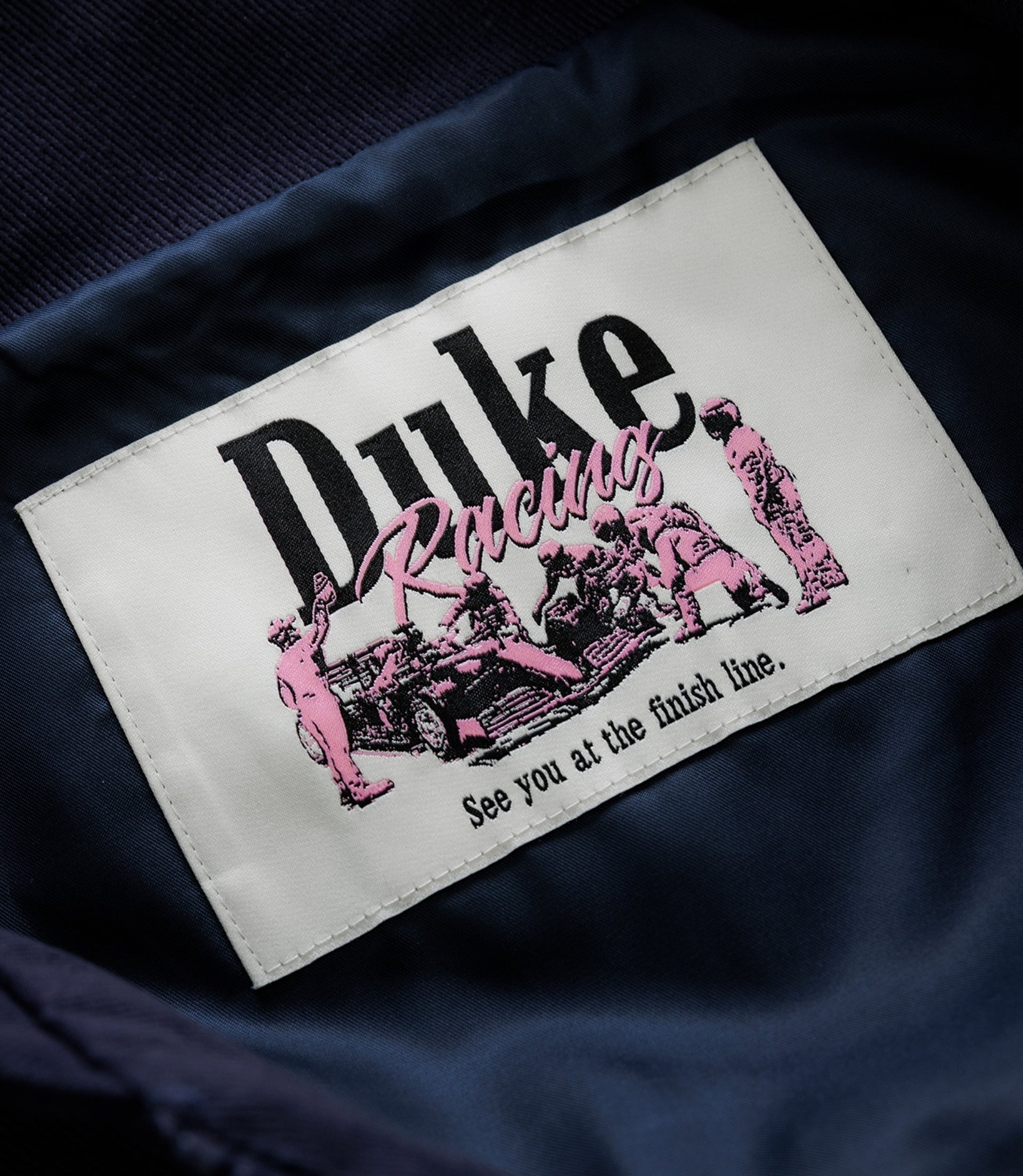 Close up of a white patch on navy fabric showing Duke Racing with pink script and racing figures