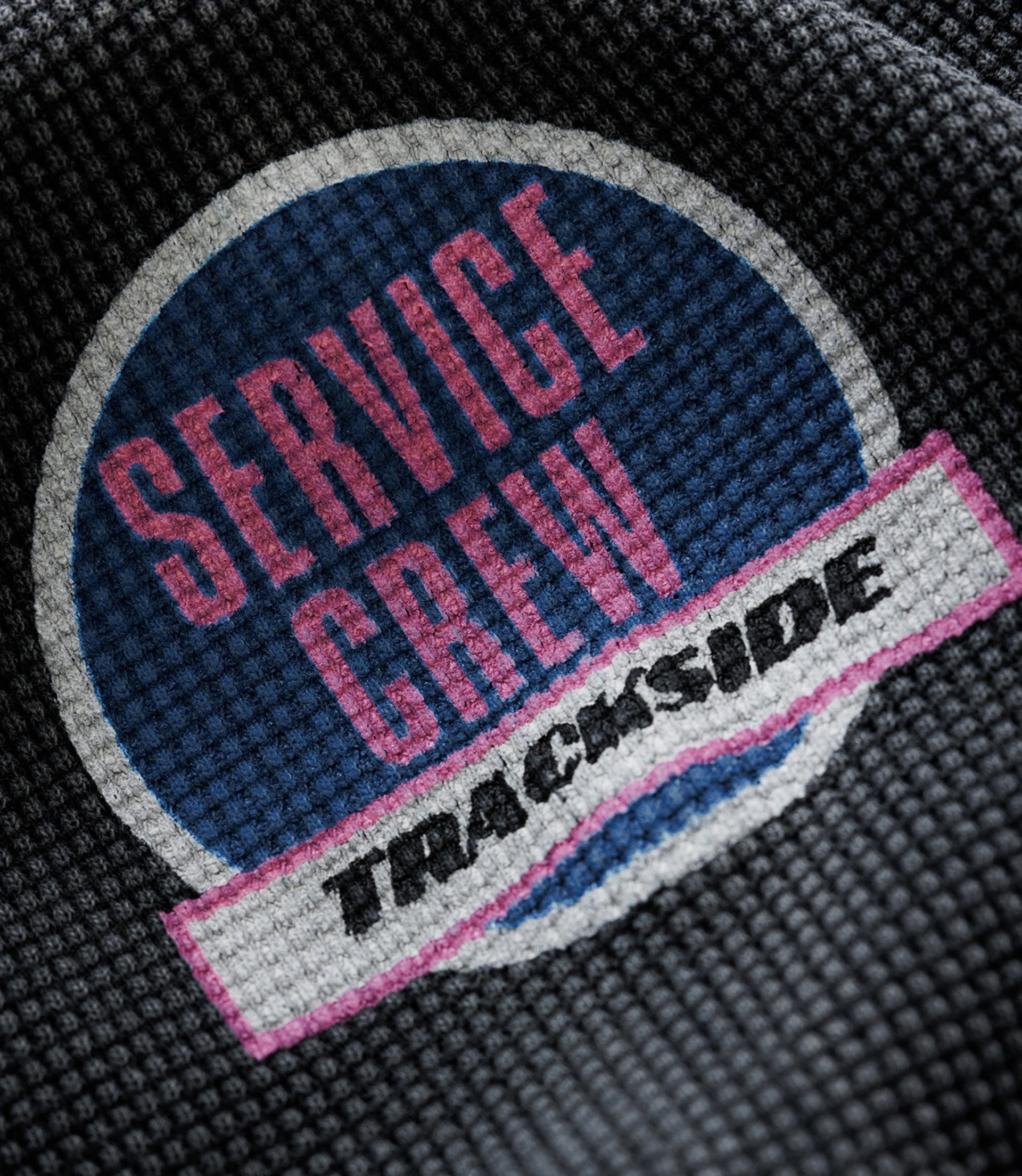 Close-up of circular patch on black washed waffle jersey featuring a blue center and pink lettering